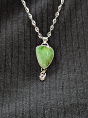 Silver Pendant and Chain with a Green Royston turquoise stone that is an angular shape and a small star dangling element at the bottom with a pink sapphire in the center. Being worn with a black striped shirt as the background.