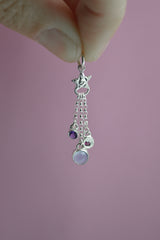 Silver charm featuring a small silver star with 3 separate chain pieces hanging from the bottom. Each has a different gemstone at the end. Purple amethyst, blue chalcedony and berry colored rhodolite garnet. Being held by fingers with a pink background.