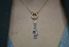 Silver charm featuring a small silver star with 3 separate chain pieces hanging from the bottom. Each has a different gemstone at the end. Purple amethyst, blue chalcedony and berry colored rhodolite garnet. Being worn on a gold chain on a circular charm holder.