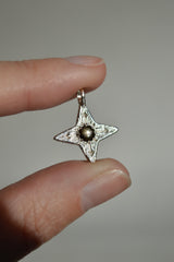 Silver Compass Star Pendant with textured surface and arrows. In the center is a black star sapphire. The "star" appears in direct sunlight as shown. Being held by fingertips.